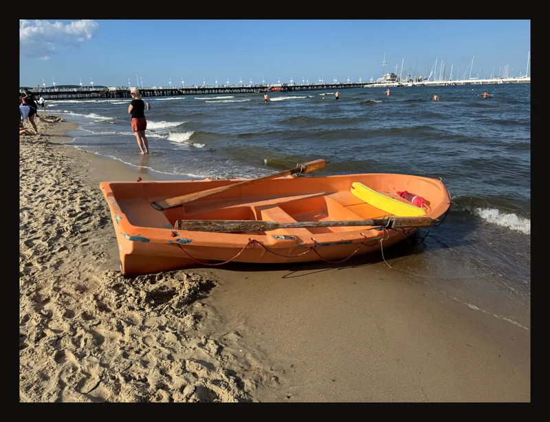 Photograph of an orange boat on a beach, with water, a pier and sky